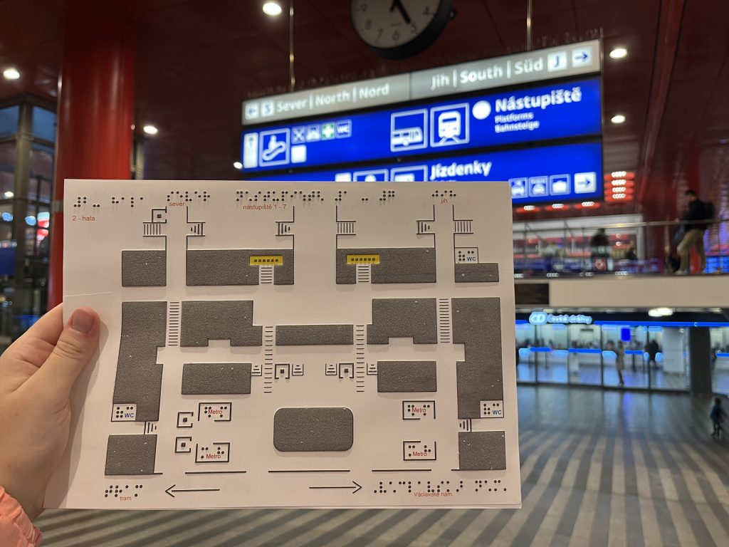 Druhý grafický list hmatáku č. 16 Praha hlavní nádraží, na pozadí odjezdová hala a prodejna lístků ČD.